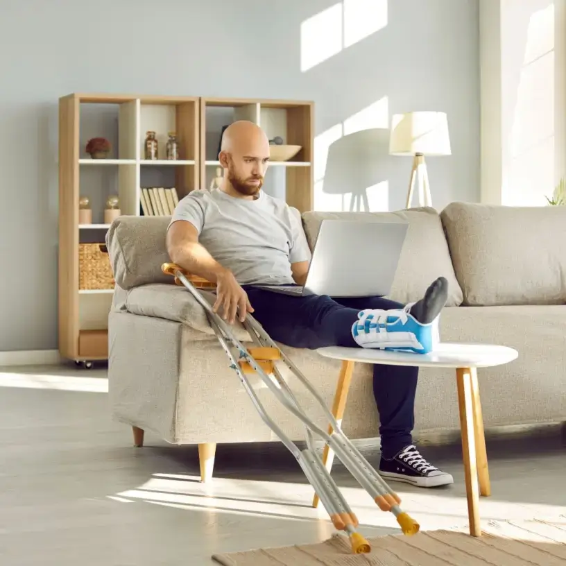 Man with crutches sits on couch working on laptop