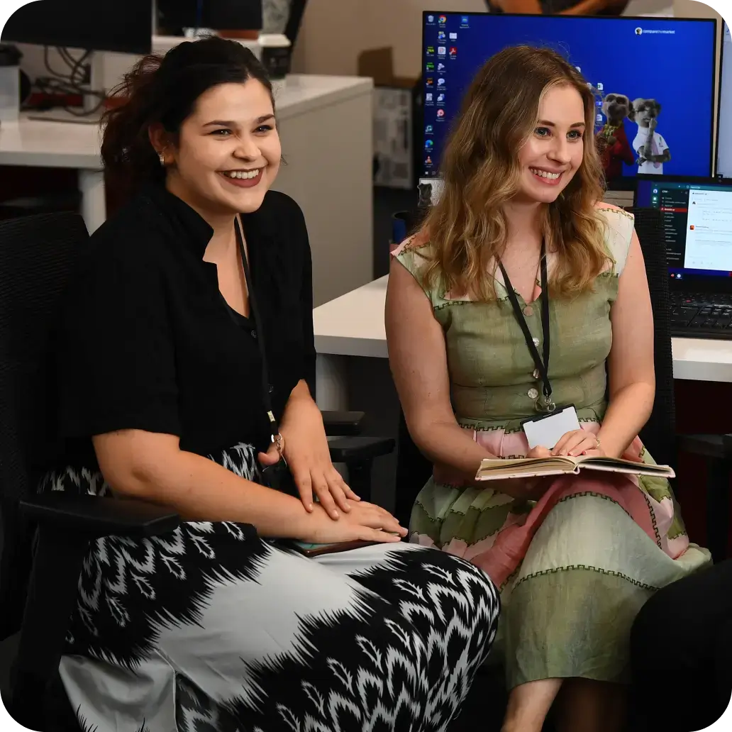 Two young women who are Compare the Market employees, sitting at a desk smiling.
